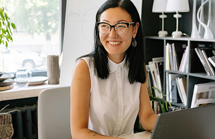 A woman wearing glasses and a sleeveless blouse sits at a desk with a laptop. A smartphone, a small plant, and a flip chart are also on or near the desk. Shelves with books are in the background.