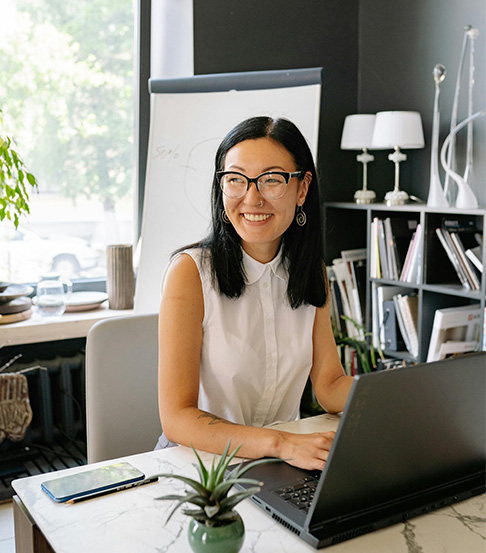 A woman wearing glasses and a sleeveless blouse sits at a desk with a laptop. A smartphone, a small plant, and a flip chart are also on or near the desk. Shelves with books are in the background.